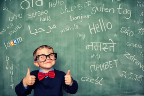 boy wearing glasses with thumbs up and hello written in multiple languages on chalkboard